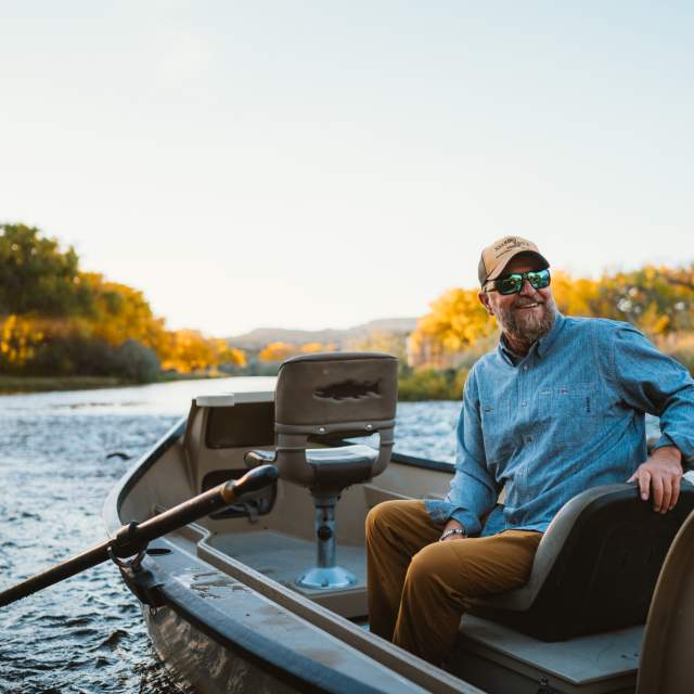 A man in a blue shirt, brown pants, a hat and sunglasses sits on a small fishing boat with oars in the water of the river surrounded by banks with golden plants. he smiles looking back past the camera