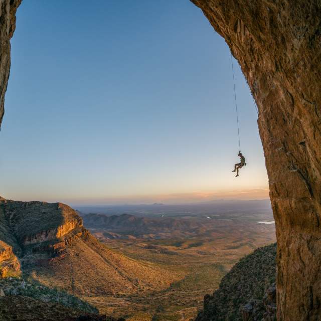 Climber Ben Hanna hangs from a rope anchored into a large arch, overlooking a horizon featuring cliffs and plains