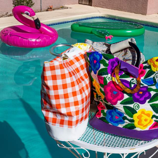A colorful poolside scene featuring a vibrant, floral-patterned bag and a checked towel on a white metal table, with inflatable pool rings floating in the background.