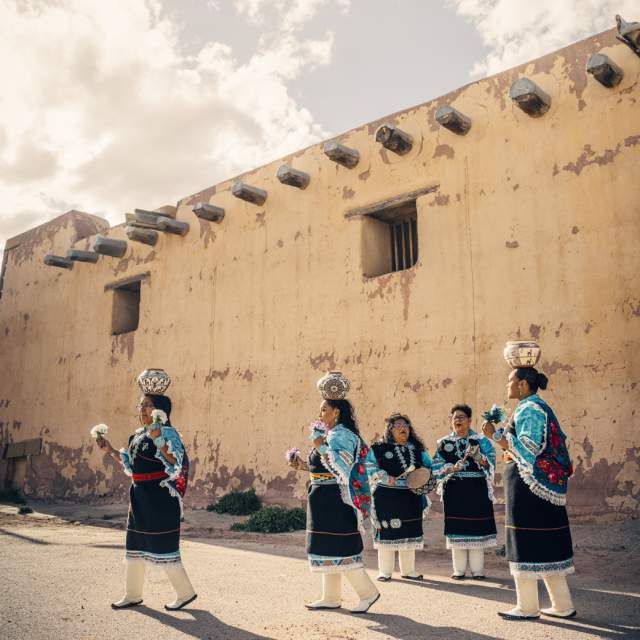 Traditional Zuni dancers in black, blue, pink and white traditional garb dance with instruments and pottery in front of a weathered adobe wall