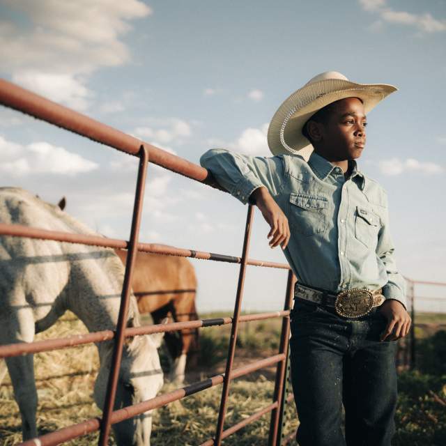 Young person in a cowboy hat and denim shirt leaning on a fence with horses behind it in a pasture.