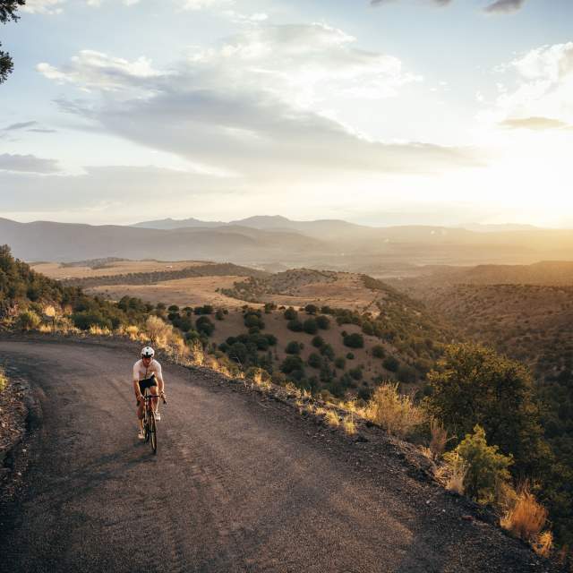 A person rides a bicycle on a mountainous road during sunset. The landscape features rocky cliffs, rolling hills, and a vast, scenic view under a partly cloudy sky.