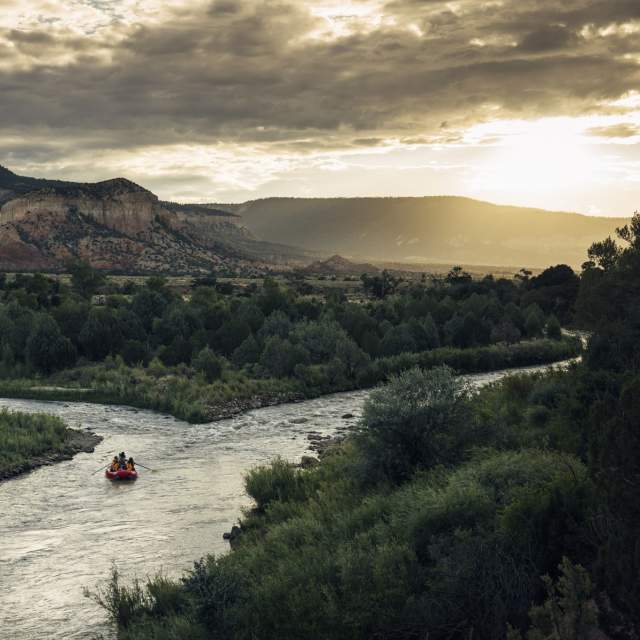 a small red raft with two people floats to a split in a river with the sun peeking over the mountains in the background
