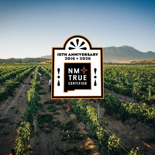 A vast vineyard stretches under a clear sky, with mountains in the background. In the center, there's a large "NM True Certified" logo celebrating its 10th anniversary from 2016 to 2026.