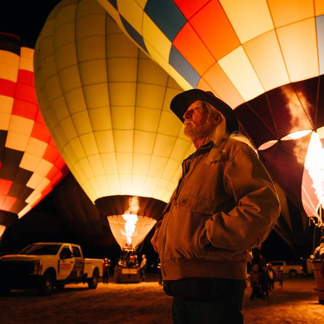 A person wearing a hat stands in front of several illuminated hot air balloons at night. The flames from the burners light up the balloons, creating a colorful display. The scene includes a vehicle in the background among the balloons.