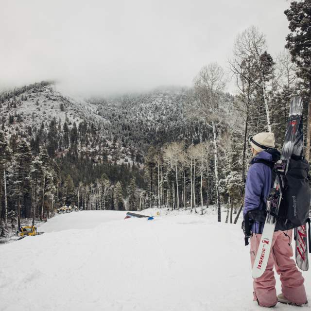 A person wearing winter clothing stands on a snowy slope, holding skis branded with "Line." They are looking towards a snow-dusted mountain landscape with tall trees around. Some snow-covered structures and ski equipment are visible in the distance.