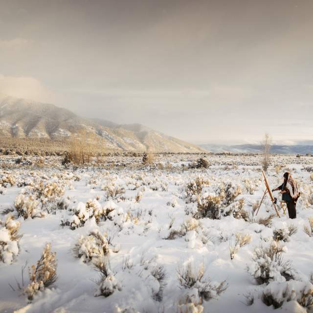 A person stands in a snow-covered landscape in front of a canvas, surrounded by sparse trees and bushes. In the background, there are snow-capped mountains under a cloudy sky.