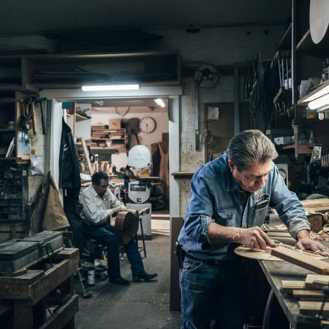 A person is working intently on a piece of wood in a cluttered workshop, while another person sits in the background holding the body of a guitar.
