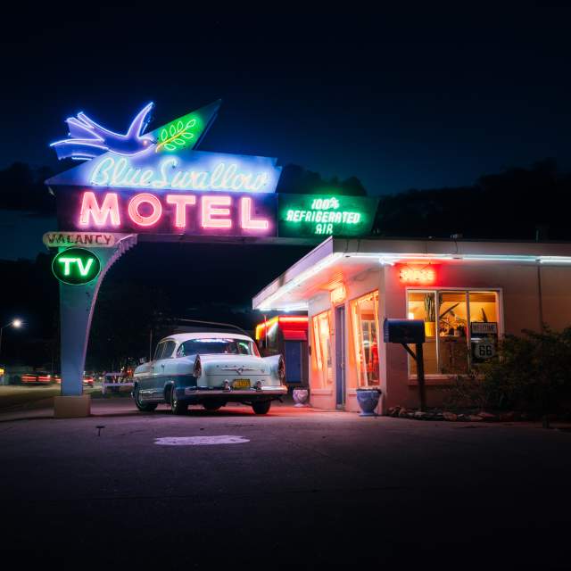 Night view of the Blue Swallow Motel with vibrant neon signs, featuring a classic car parked in front.