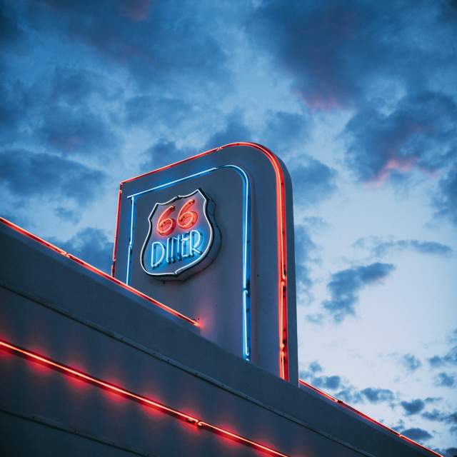 Neon sign for '66 Diner' illuminated in blue and red against a twilight sky with clouds.