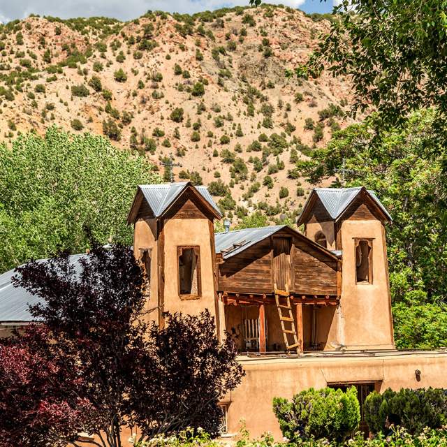 Traditional adobe church nestled against a hill covered with green vegetation, under a partly cloudy sky.