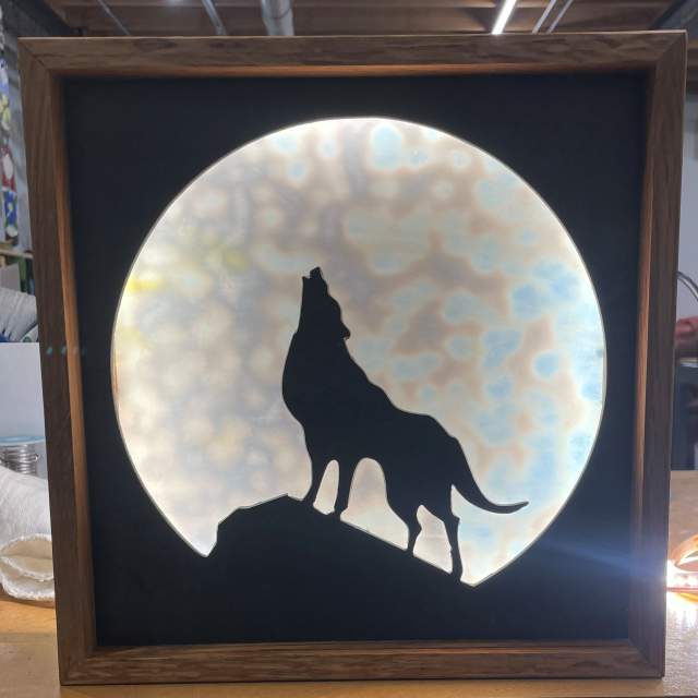 Stained glass silhouette of a wolf howling on a hill against a luminous, textured backdrop, framed in a dark square frame.