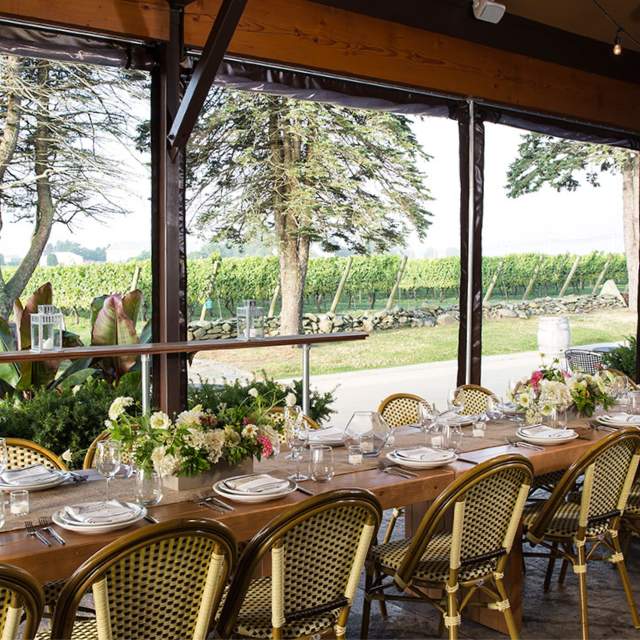 A dining table set with plates, glasses, and flowers in a room with large windows overlooking a vineyard.