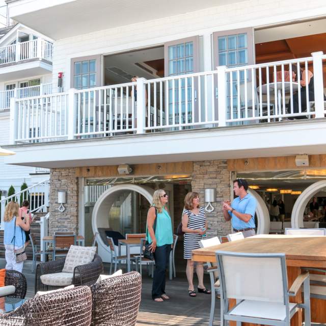 A group of people socializing on the outdoor patio of a building with a balcony and stone facade.