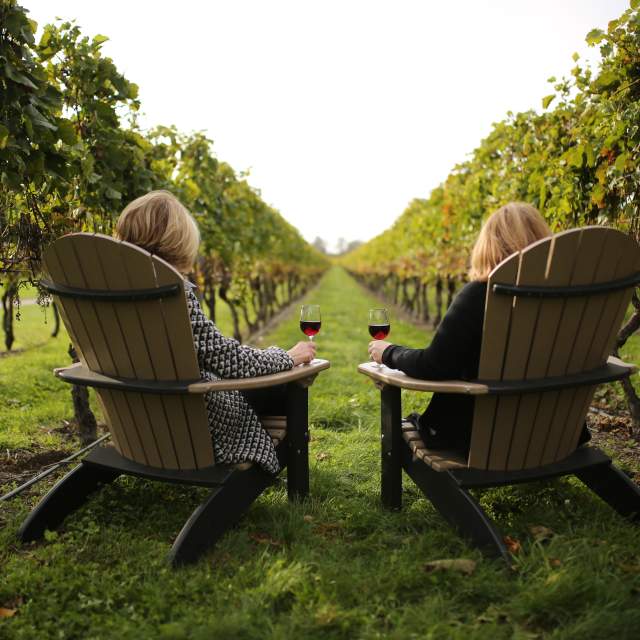 Two women sitting in Adirondack chairs in a vineyard with wine glasses