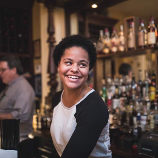 Young Black woman smiling with the bourbon collection of Tousey House Tavern behind her