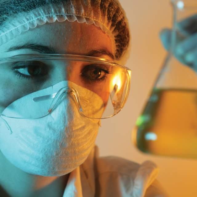 Woman wearing mask and goggles and looking at a beaker of yellow liquid.