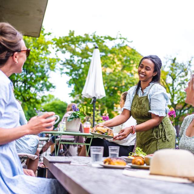 En unge dame serverer to damer mat på uteserveringen til fru Blom