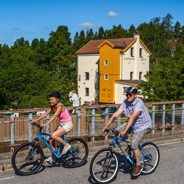 Ett par på sykkeltur over brua foran Strømsfoss mølle