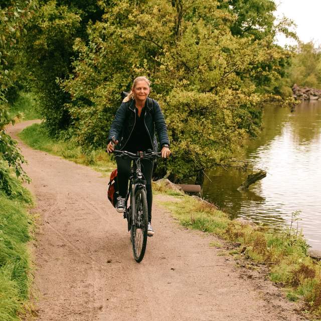 Kvinne som sykler langs en gruslagt vei på Glommastien, med Glomma i bakgrunnen.