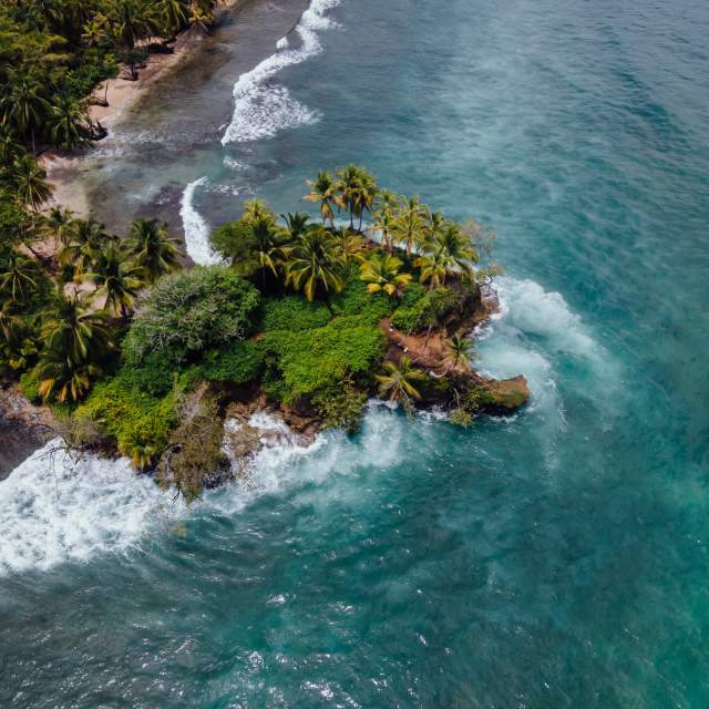 : Aerial view of a small tropical island with lush vegetation, a white sand beach, and surrounding turquoise waters with coral reefs and gentle waves