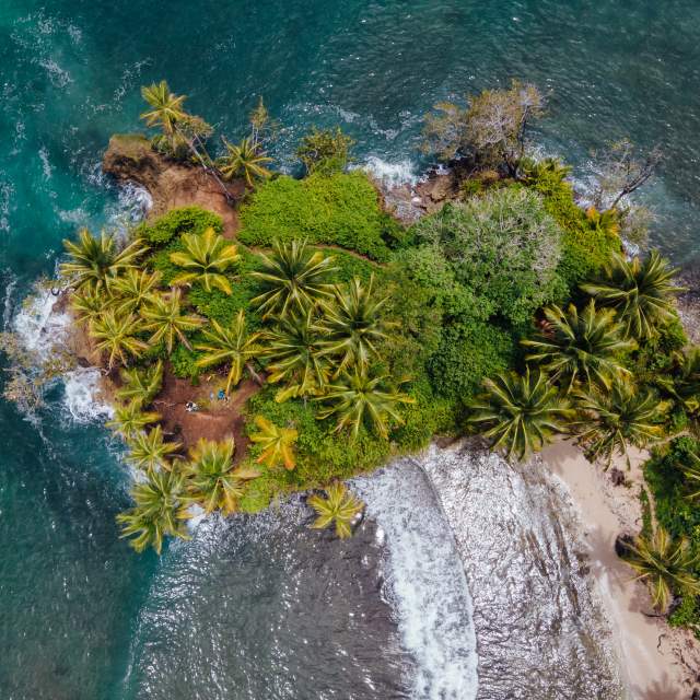 Aerial view of a small tropical island with lush vegetation, a white sand beach, and surrounding turquoise waters with coral reefs and gentle waves