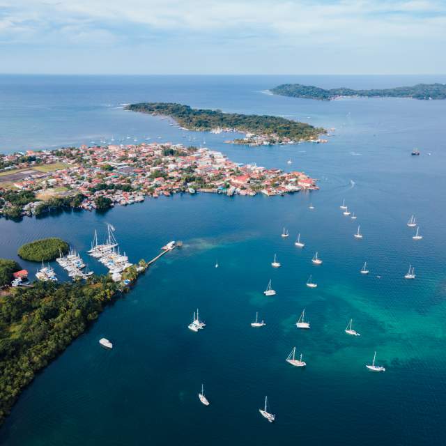 An aerial view shows a vibrant coastal town with colorful, red-roofed buildings situated among lush green islands and a tranquil bay filled with sailboats. A marina and a small airport are also visible, suggesting a popular sailing destination.