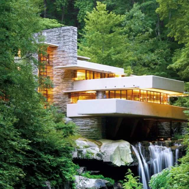 A modern home with a balcony and a waterfall coming out of the bottom. Green luscious trees are surrounding the architectural marvel.
