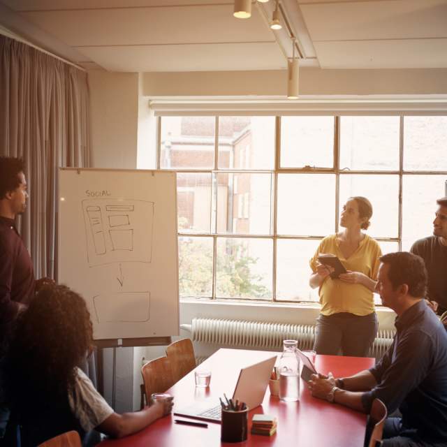 Digital Strategy as a Managed Service | Campaign Management - Woman and team of staff in an office looking at a campaign idea on a presentation easel