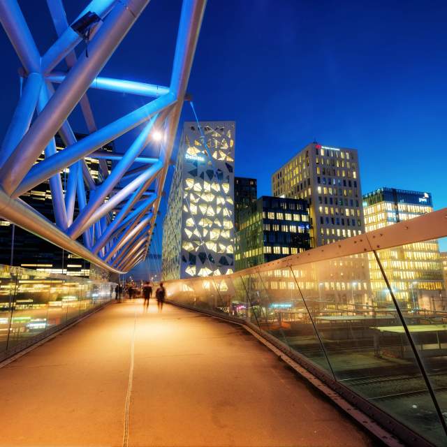 People walking across a bridge at nighttime, with a skyline in the background