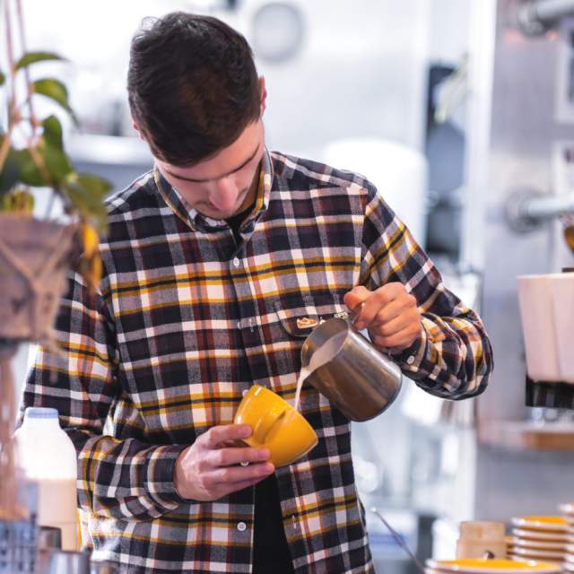 Person behind coffee bar pouring hot milk into a coffee cup
