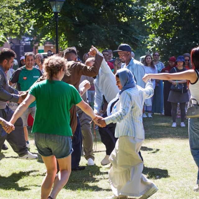 Group of people holding hands and dancing in a circle in Palmerston Park for Refugee Week