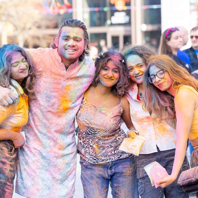 5 people with their arms around each other covered in colourful powder smiling at the Holi event in Guildhall Square