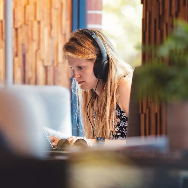 Person in background with headphones on looking at a laptop. There is a blurred plant and persons face in the foreground at room2 Southampton