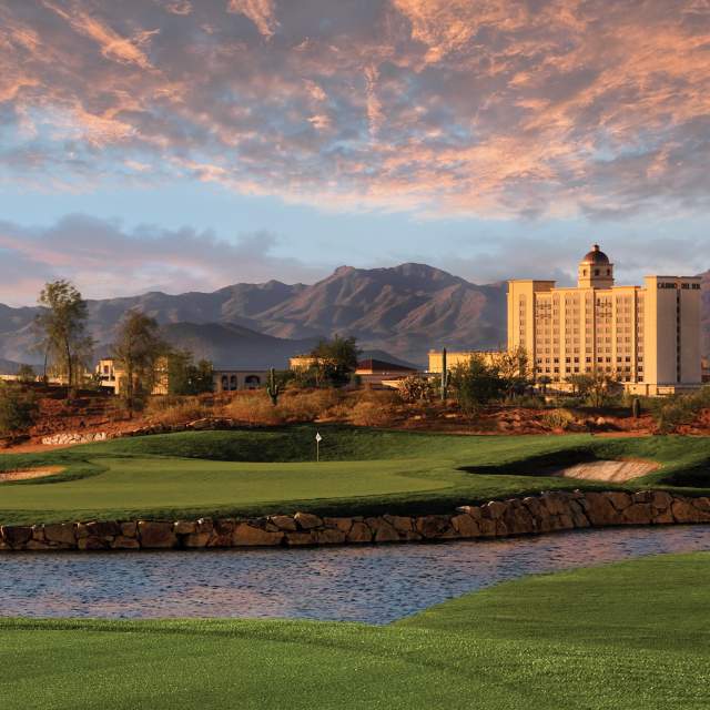 Exterior of Casino Del Sol, Wide shot showcasing the green landscape and pond of the Sewailo Golf Course. Casino Del Sol stands tall and golden against the desert mountains