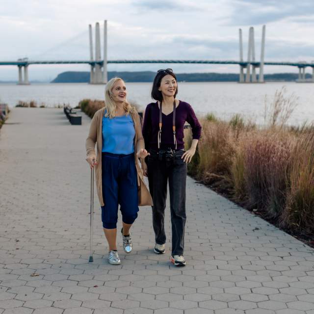 Two people are walking along the Riverwalk in Tarrytown. The Gov. Mario Cuomo Bridge is in the background.