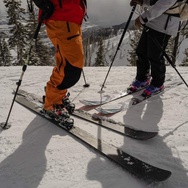 Two skiers pause on a snow-covered mountain slope near Jackson, Wyoming, with a wintry mountain landscape and evergreen trees in the background. The close-up view of skis, poles and winter gear highlights downhill skiing and outdoor recreation in Teton County.