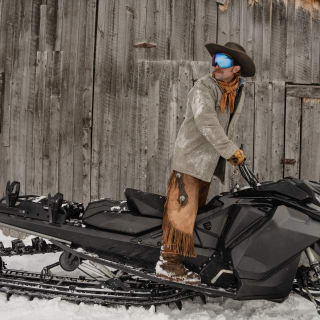A cowboy dressed in winter Western attire stands on a snowmobile beside a weathered wooden barn near Jackson, Wyoming. Snow-covered chaps, a cowboy hat and ski goggles blend Wyoming’s Western heritage with modern winter recreation in Teton County.