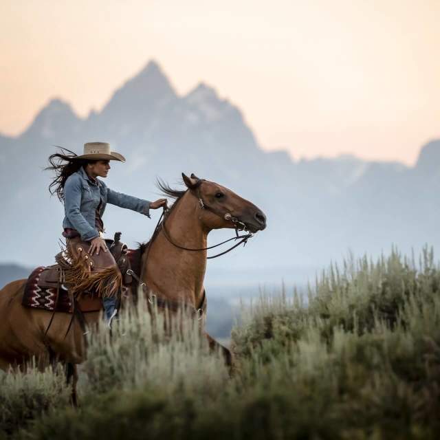 Woman riding her horse up a hill with the Grand Tetons in the background.