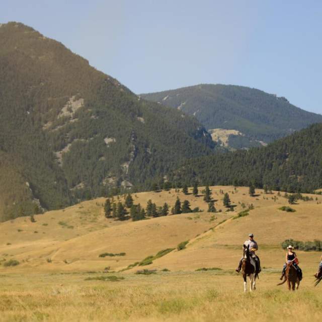 Horseback riders in grassy field with mountain ranges in the background.