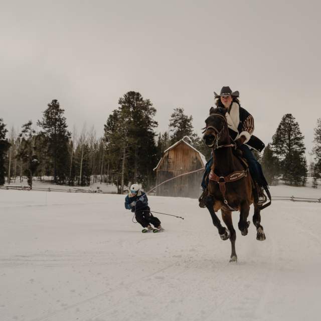 A horseback rider pulls a skier through the snow during a skijoring run near Wyoming. The horse gallops across a snow-covered field as the skier follows behind, with a rustic wooden barn, fencing, and evergreen forest creating a classic Wyoming winter setting.