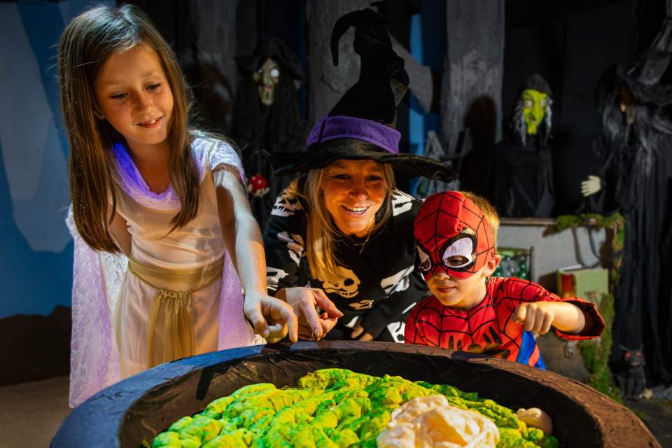 A group of children in costumes smile and look into a cauldron full of green potion.