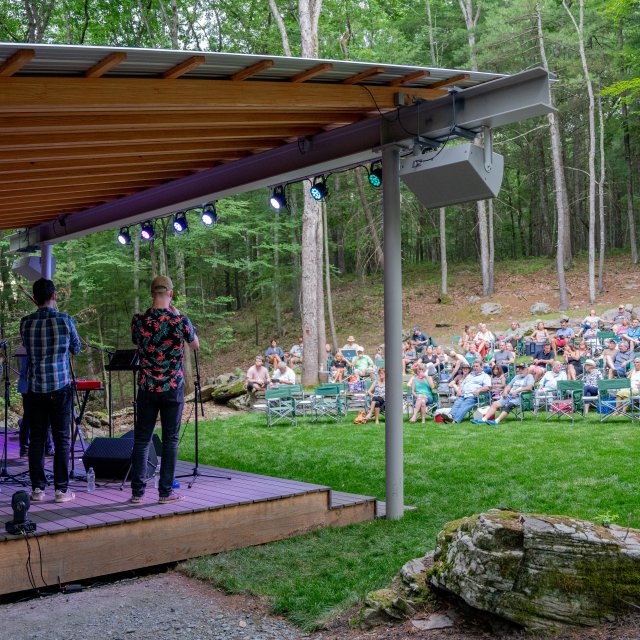 A band performs for a crowd on an outdoor stage surrounded by woods.