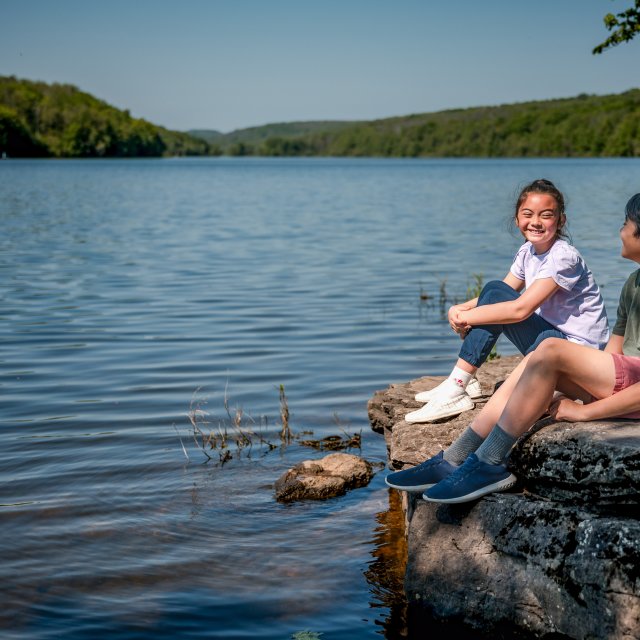 Two children smiling while sitting on a rock in front of a lake during the summer.