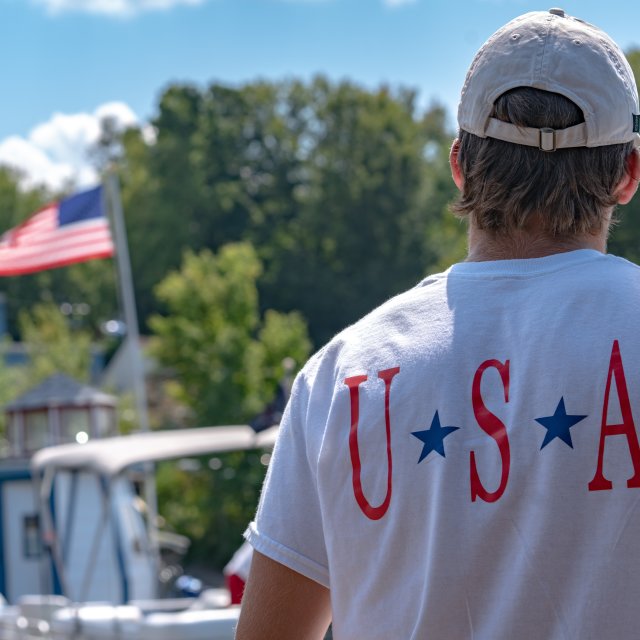 A man wearing a USA shirt looks at an American flag at a lake in the Poconos.