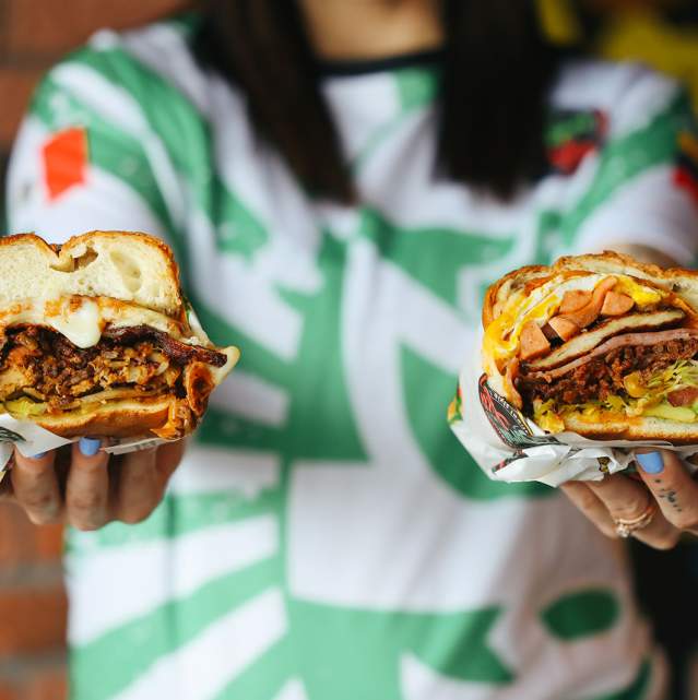 A woman in a green and white shirt holding two torta sandwiches out to the camera