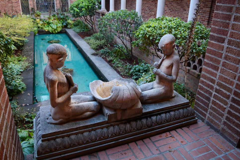 A statue of two people seated across from each other and gazing at a large beautiful shell in between them. A tiled pond and trees are in the background.