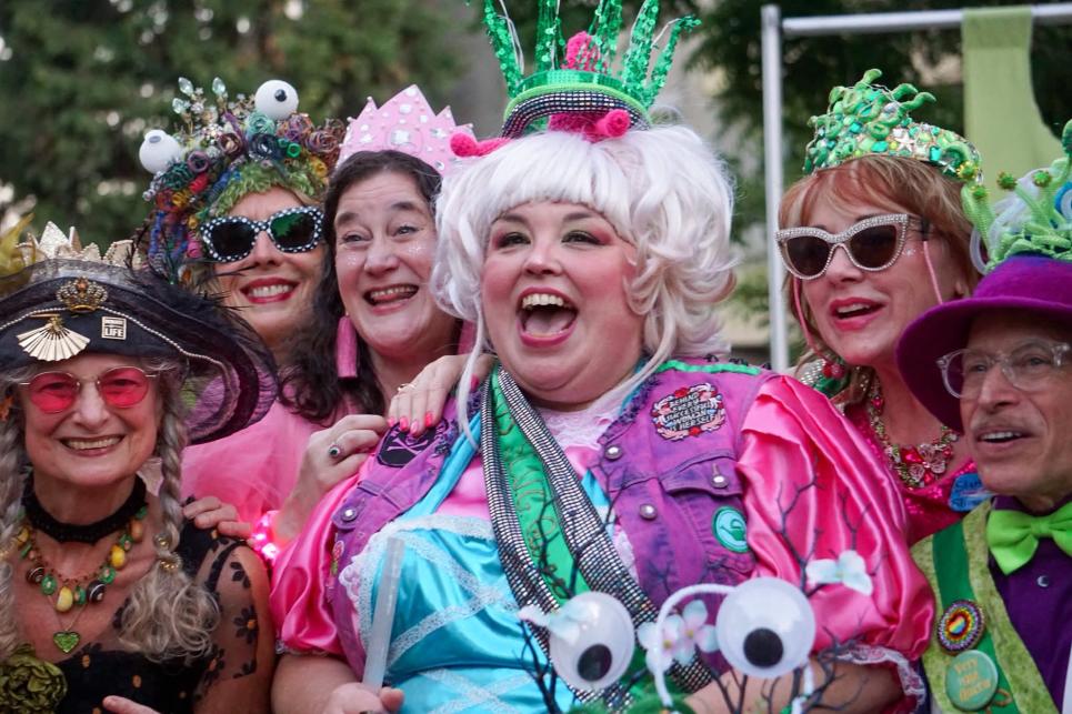 A person in a white wig and colorful clothes with a green crown is standing with the other dressed up people.