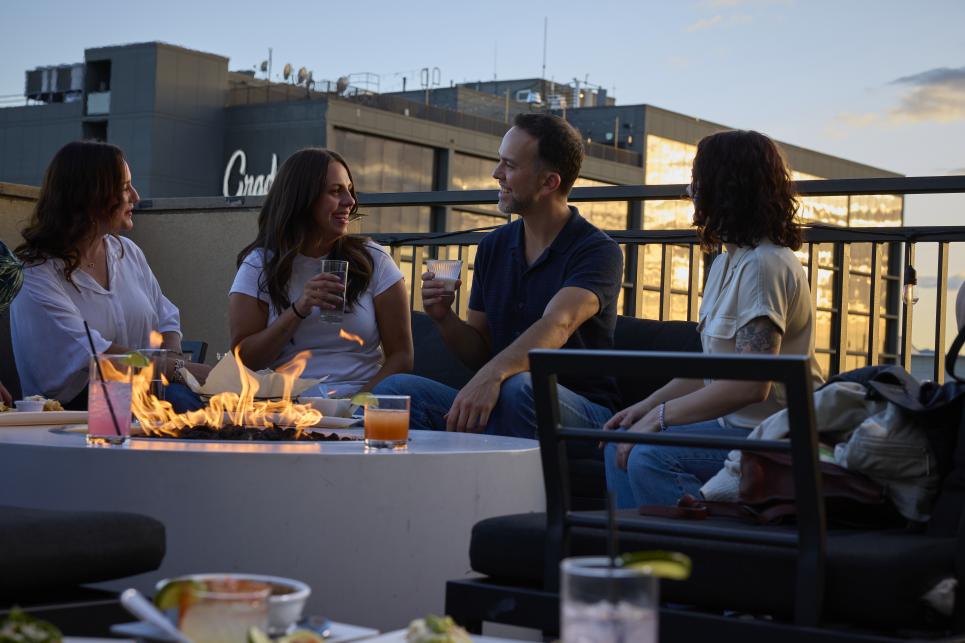 People sitting outside on a rooftop deck around a fire with cocktails.
