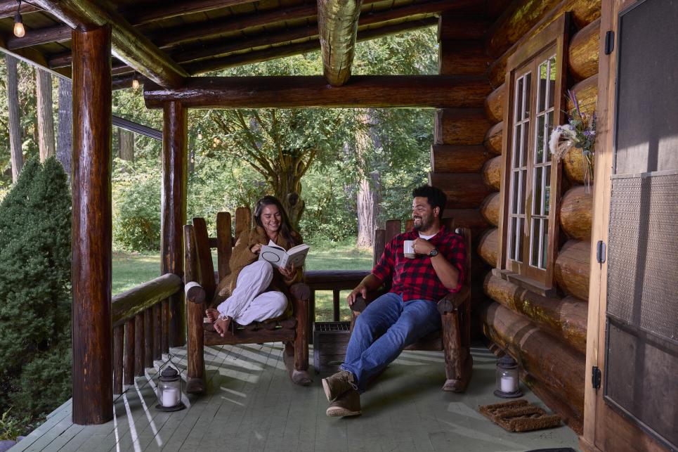 Two people sit on the porch of the log lodge. One reads a book, the other sips a warm beverage.